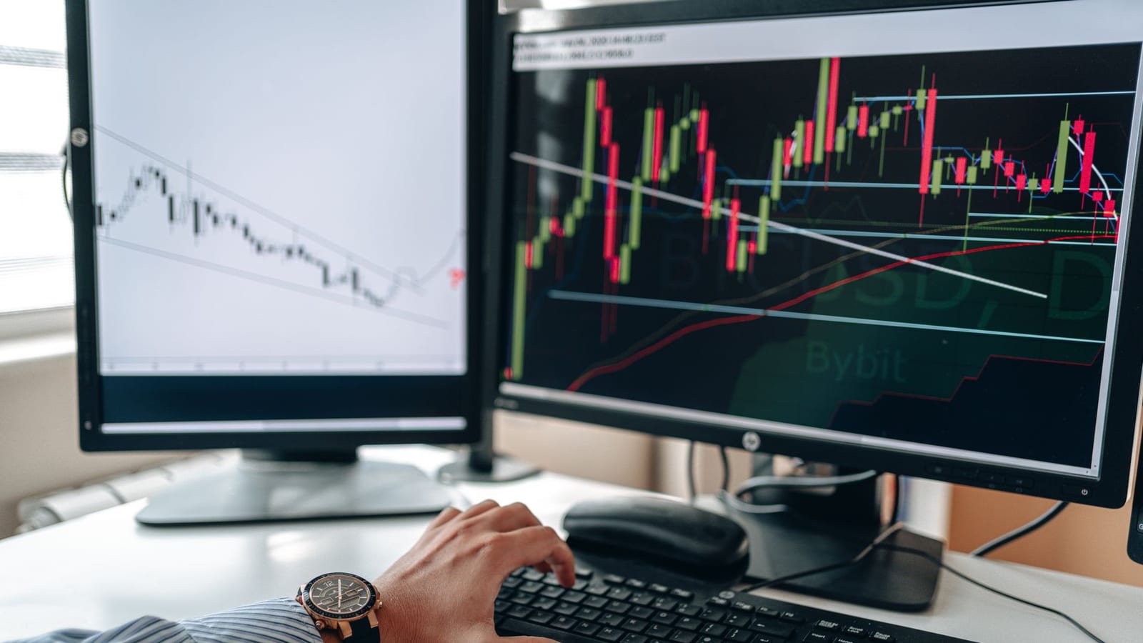 Duas telas com gráficos de candlestick e mão no teclado