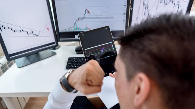 Trader analisando gráficos em tablet e três monitores