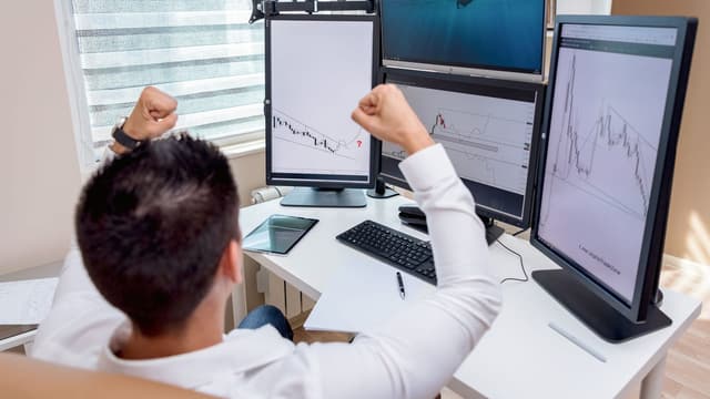 Trader com os braços erguidos em frente a quatro telas de operação